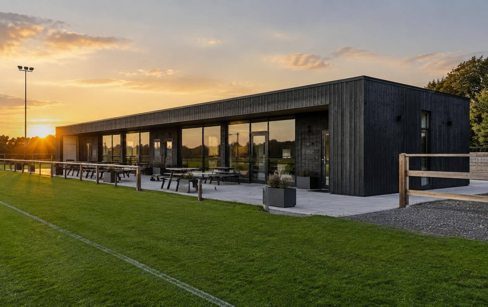 Modern dark-clad modular clubhouse on a sports ground at sunset
